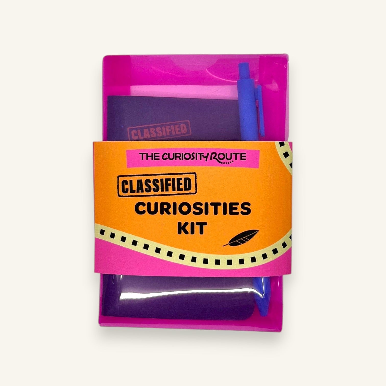 The Curiosity Route Classified Curiosities Kit in a pink envelope with a blue pen and notebook for secret messages.