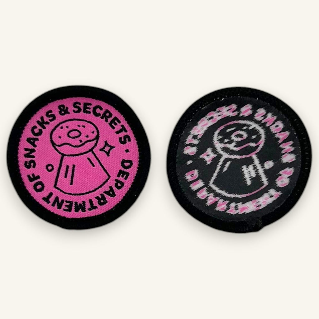 The Curiosity Route circular embroidered patches in pink and black featuring Department of Snacks and Secrets text